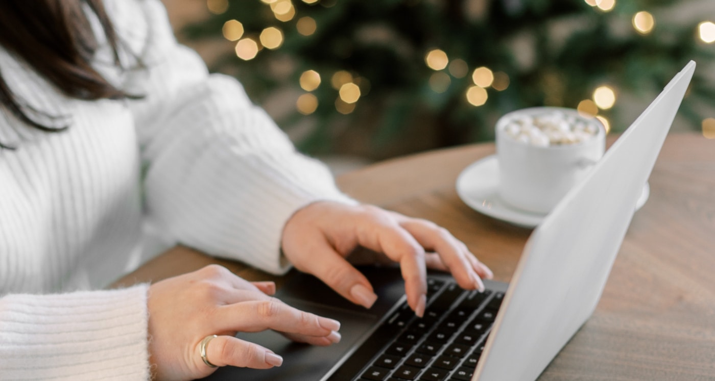 woman working on laptop beside holiday tree learning How To Create A Christmas Budget And Stick To It