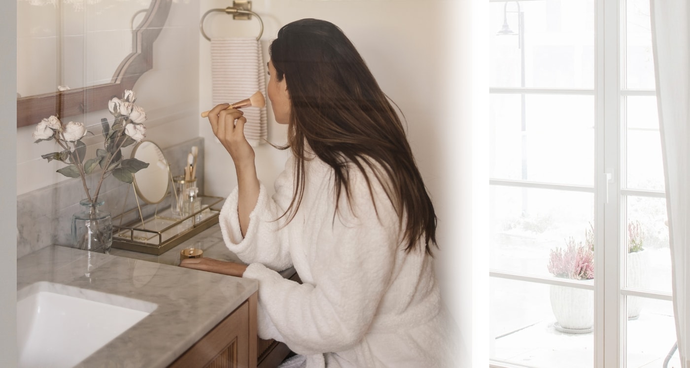 woman applying makeup in her bathroom while reading about How To Spend Less Money On Makeup